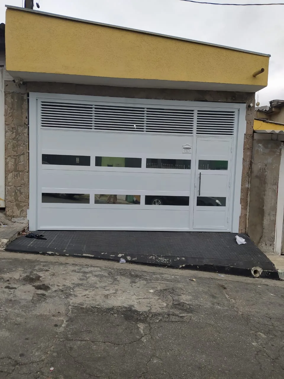 Portão de garagem branco com veneziana e vidro em fachada amarela